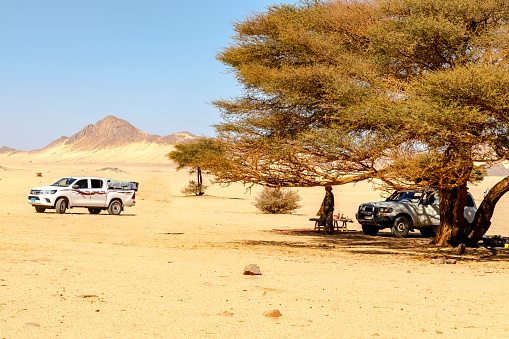 Djanet, Algeria -DECEMBER 19, 2021: unrecognizable tuareg man, food table under Acacia tree . Dry herbs, two off-road cars, yellow coloured sand stones, rocky mountain blue sky