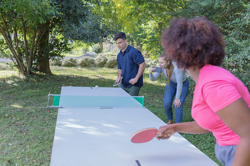A male and a black female business executives  are playing table tennis at a park outside their office while a female executive standing near the net happily records  with her smartphone