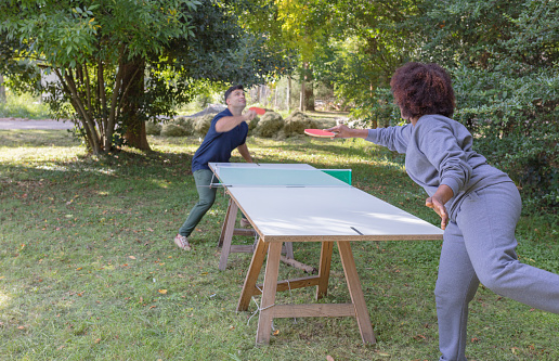 A black female business executive  is playing ping-pong with a male co-worker at park outside office. She is hitting the ball.