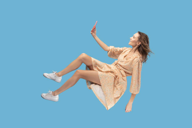 happy woman levitating with mobile phone, reading message chatting happy in social network online Hovering in air. Smiling girl in yellow dress levitating with mobile phone, reading message chatting happy joyful in social network online, surfing web while flying. indoor shot isolated on blue zero gravity stock pictures, royalty-free photos & images