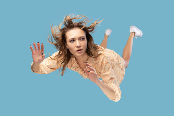 Beautiful young woman levitating in mid-air, falling down and her hair messed up soaring from wind Beautiful young woman levitating in mid-air, falling down and her hair messed up soaring from wind, model flying hovering with dreamy peaceful expression. indoor shot isolated on blue background zero gravity stock pictures, royalty-free photos & images