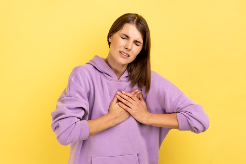 Portrait of overworked depressed woman frowning suffering sudden heart attack, myocardial infarction, risk of breast cancer, wearing purple hoodie. Indoor studio shot isolated on yellow background.