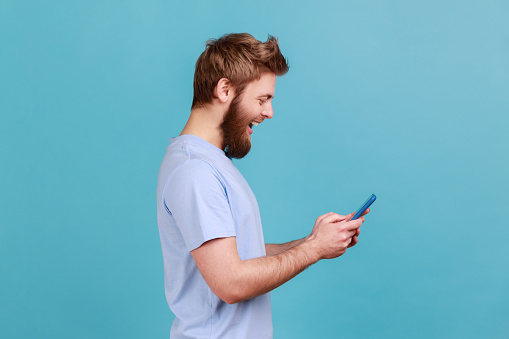 Side view of happy excited bearded man using online service on mobile phone and expressing pleasant emotions, texting messages for friends. Indoor studio shot isolated on blue background.