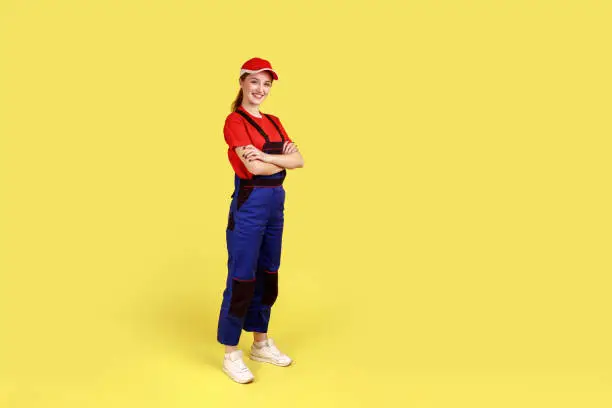 Full length portrait of happy smiling handy woman standing with crossed hands, having positive confident expression, wearing overalls and red cap. Indoor studio shot isolated on yellow background.