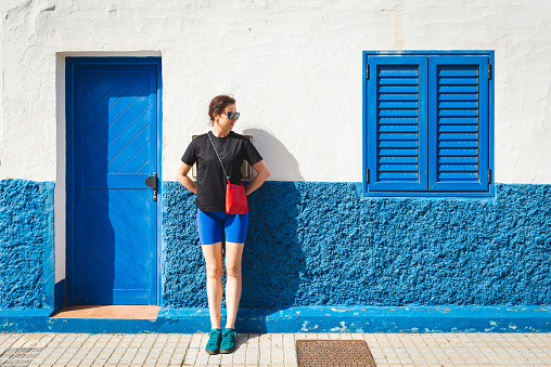 Beautiful young female standing by the wall of a blue/white building wall. Hands behind back, looking aside.