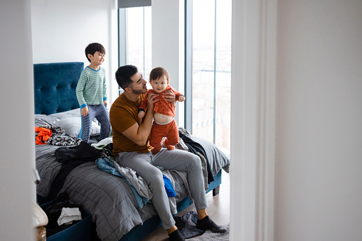 An Asian man and his two young sons wearing casual clothing and pyjamas in his bedroom on a sunny winters morning. He is getting his baby boy ready as his other son is having fun bouncing on the bed.