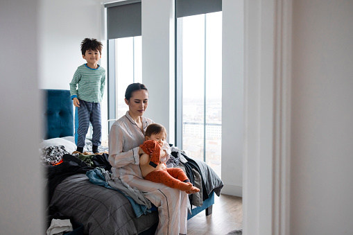 An Asian woman and her two young sons wearing pyjamas in her bedroom on a sunny winters morning. She is getting her baby boy ready as her other son is having fun bouncing on the bed.