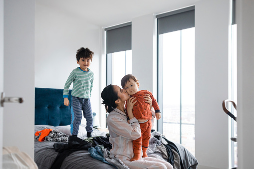 An Asian woman and her two young sons wearing pyjamas in her bedroom on a sunny winters morning. She is getting her baby boy ready and kissing him on the cheek as her other son is having fun bouncing on the bed.
