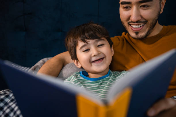I Love This Story An Asian man and his young son wearing pyjamas in his bedroom on a winter's night. They are lying in bed while the father reads his son a bedtime story. single father stock pictures, royalty-free photos & images