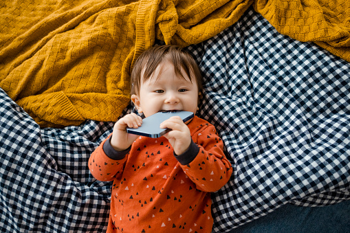 A high angle view of an Asian baby boy wearing pyjamas in a bedroom on a sunny winters morning. He is lying on a bed as he holds a smartphone in his mouth.