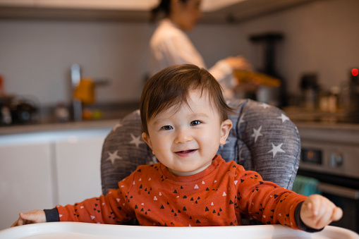 A portrait shot of an Asian baby boy wearing pyjamas on a sunny winters morning. He is sitting in a highchair in the kitchen as his mother is making his breakfast.