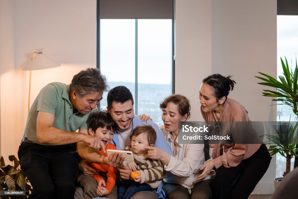 That's a Great One! A multi-generation family of six wearing casual clothing in a living room. They are sitting on a sofa and looking at a selfie they have taken on a smartphone. 30-34 Years Stock Photo