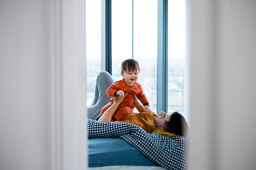 An Asian man and his baby boy wearing pyjamas in his bedroom on a sunny winters morning. He is lying down on the bed and holding his son as they spend quality time together.