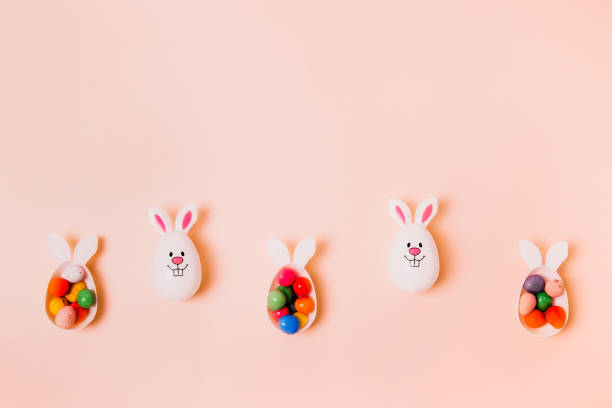 istock Top view festive Easter beige peach color background with funny bunny eggs containers filled with candy chocolate eggs. Bunny rabbit symbol. Easter egg hunt. Creative Easter. Flat lay. Copy space. 1383910564