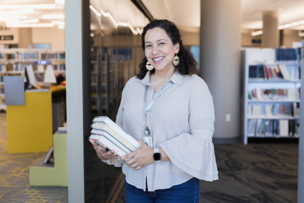Dedicated female librarian carries books to re-shelve A portrait of a smiling, dedicated mid adult female librarian carrying a pile of books to re-shelve. librarian stock pictures, royalty-free photos & images