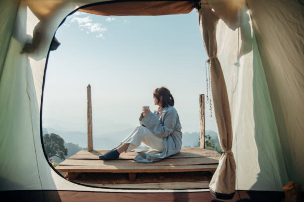 Rearview image of a woman sitting on a wooden balcony while watching a beautiful mountain and nature view outside the tent with her cup of coffee. stock photo