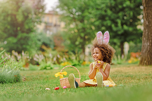 Little Black girl wear bunny ears and gathering Easter eggs on Easter egg hunt in garden