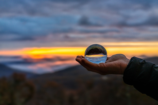 Hand holding crystal glass lens ball in palm view of round globe with reflection of fog mist clouds over blue ridge mountains at colorful sunrise in Wintergreen Resort, Virginia