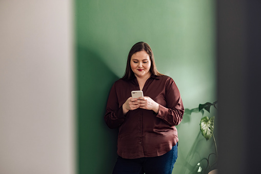 Beautiful plus size woman leaning against a green wall, smiling, looking down at her mobile phone and texting.