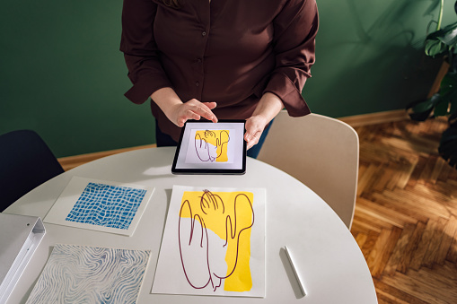 High angle view of unrecognizable overweight woman standing by the table with digital tablet while holding and comparing her illustrations. Whole screen is visible.