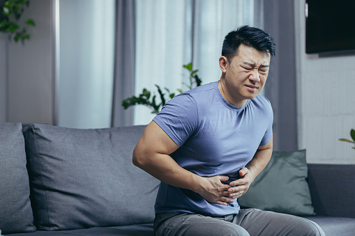 Hombre asiático en casa enfermo sentado en el sofá, tiene dolor abdominal severo, hombre en la sala de estar photo