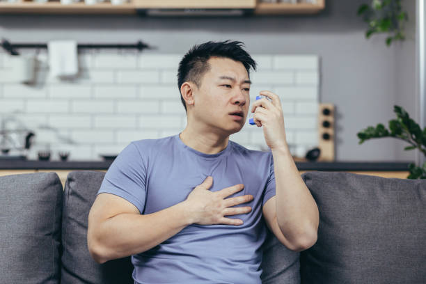 A man with asthma, an Asian man at home sitting on the couch breathing in an inhaler A man with asthma, an Asian man at home sitting on the couch breathing in an inhaler asthma stock pictures, royalty-free photos & images