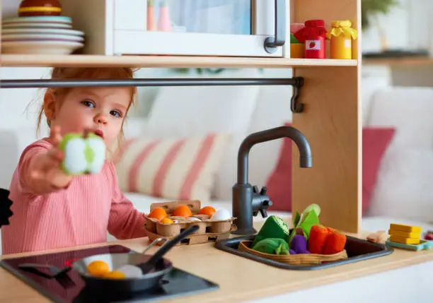 Photo of cute toddler baby girl playing on toy kitchen at home, roasting eggs and treat you with apple slice, let's share