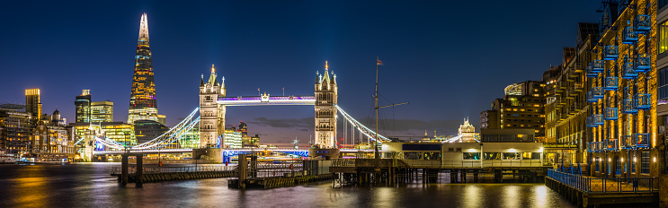 Panoramic view across the River Thames to the historic span of Tower Bridge illuminated against the blue dusk sky overlooked by the futuristic glass spire of The Shard in the heart of London, Britain's vibrant capital city.