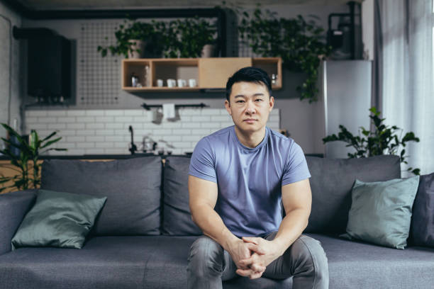 portrait d’un homme sérieux à la maison, homme asiatique assis sur le canapé regardant attentivement la caméra - domestic car audio photos et images de collection