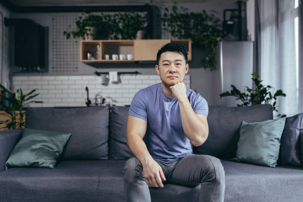 portrait d’un homme sérieux à la maison, homme asiatique assis sur le canapé regardant attentivement la caméra - domestic car audio photos et images de collection