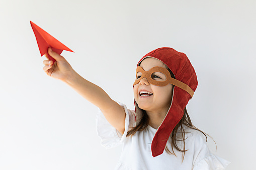 Little girl  is holding model paper plane in hand wearing pilot costume smiling and looking at it in front of white background.