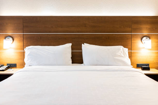 istock Bedroom empty room with illuminated lit lamp light and front view of white pillows with sheets on mattress bed headboard in modern hotel with nobody 1384045494