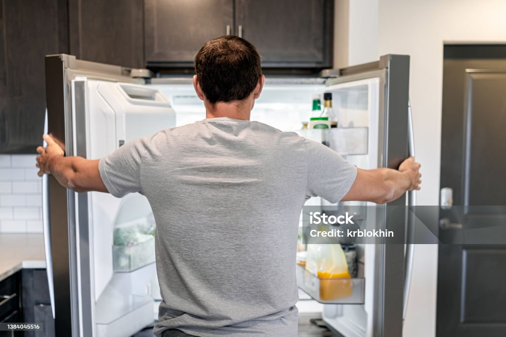 Back of hungry man opening fridge refrigerator doors domestic appliance searching for food inside with condiments and juice in modern kitchen Refrigerator Stock Photo