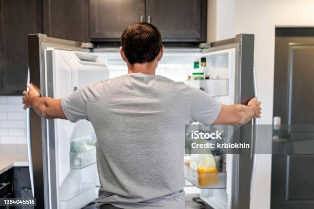 Back Of Hungry Man Opening Fridge Refrigerator Doors Domestic Appliance Searching For Food Inside With Condiments And Juice In Modern Kitchen Stock Photo - Download Image Now
