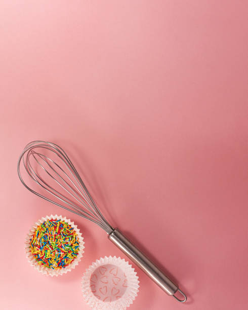 istock Cupcake liners with colored sugar inside whisk at the bottom from above flat lay top view. Confectionery cooking concept with copy space on bright pink paper background. National cooking day. 1384049560