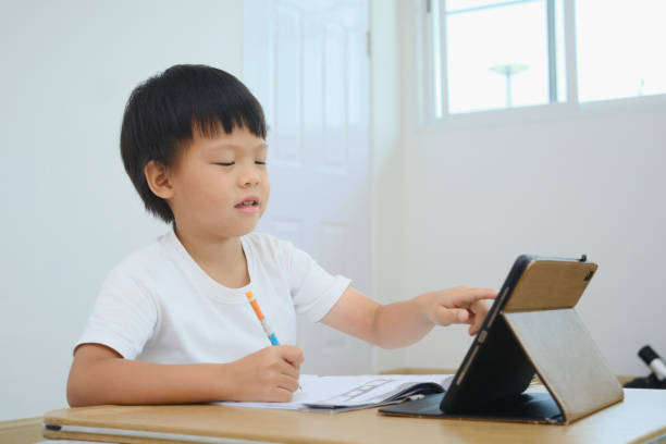 istock Asian little kindergarten kid using tablet computer, studying homework during online lesson at home 1384054161