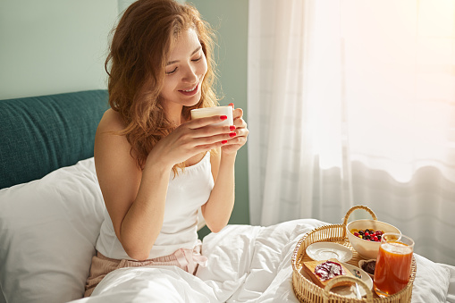 Happy young female in sleepwear drinking fresh aromatic coffee and smiling, while sitting in bed with wicker tray with delicious breakfast after awakening in sunny morning at home