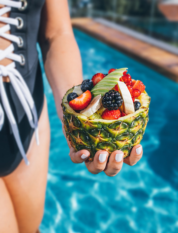 Young woman with swimwear holding a fruit bowl with various colourful wild berries, blueberry, redcurrant, strawberry, blackberry and apple in a pineapple bowl for a healthy breakfast in the swimming pool in the sunny summertime