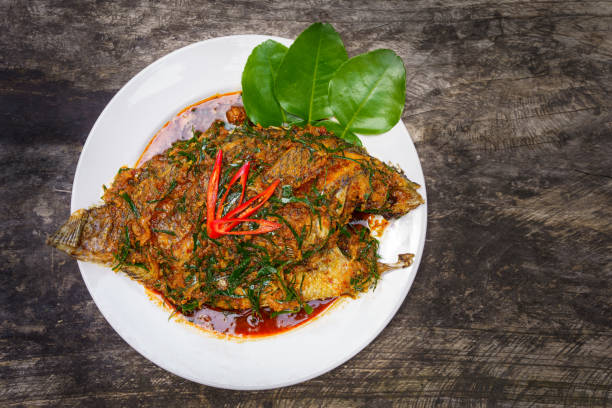 top view Fried Nile Tilapia fish with red curry paste on white plate wooden background, Thai food is spicy mixed with sweet flavor with aromatic spices, Red curry paste is popular to cook in Thailand vista superior pescado frito de tilapia del nilo con pasta de curry rojo sobre fondo de madera de plato blanco, la comida tailandesa es picante mezclada con sabor dulce con especias aromáticas, la pasta de curry rojo es popular para cocinar en tailandia