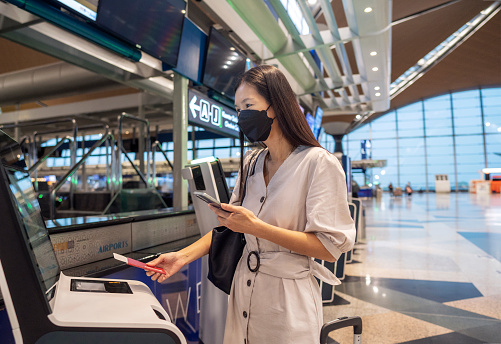 Asian bbusinesswoman at self check in kiosk in airport, holds passport, going to register for flight, wears protective face mask for safe traveling during endemic in public place