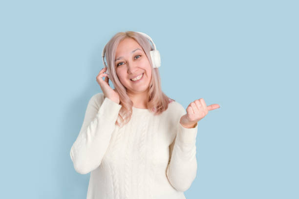 una hermosa mujer rubia de 35-40 años escucha música con auriculares inalámbricos y hace un gesto con el dedo hacia arriba - 35 40 years audio fotografías e imágenes de stock