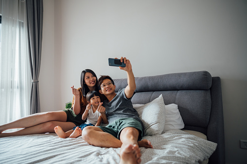 Young Asian Family lying on bed with funny facial expression and taking selfie at home.