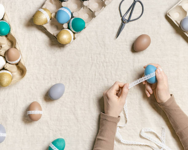 istock Decoration Easter eggs, female hands decorating colorful chicken egg with white tape lace, traditional celebration food and holiday symbol.  Home preparing for Happy Easter. Top view 1384136466