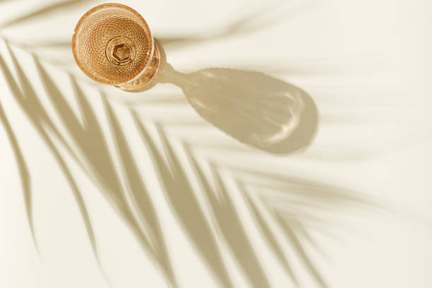 white wine glass peach color glass on beige background with palm leaf shadow, glare at sun. minimal summer rest concept. dry wine in colored glassware goblets style. creative top view - witte wijn fotos stockfoto's en -beelden