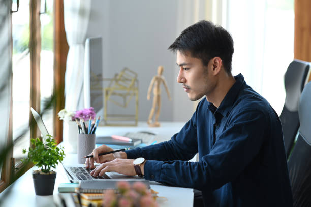 istock Focused asian man working with laptop computer at bright modern office. 1384154185