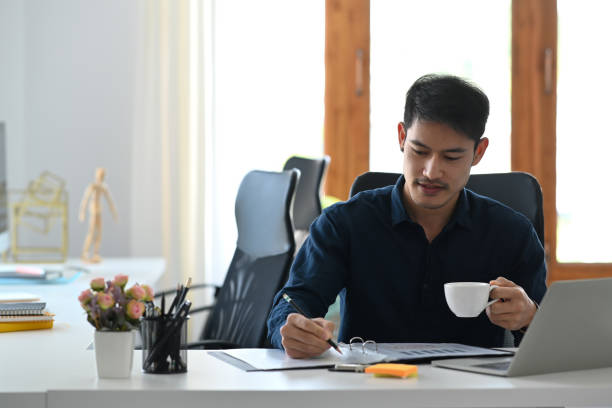 istock Asian man office worker drinking coffee and reading financial reports at workplace. 1384154188