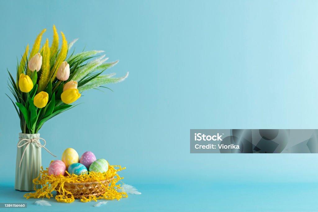 Easter eggs and tulips with space for copy Multi colored hand-painted Easter eggs in a basket with the bouquet of tulips in a vase. Blue background. Space for copy. Easter Stock Photo