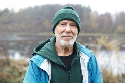 Primer plano al aire libre de un apuesto jubilado anciano activo con barba gris que camina por la mañana en la naturaleza salvaje, posando contra un lago brumoso y un colorido fondo de bosque otoñal, mirando a la cámara photo
