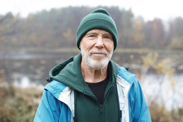 outdoor close up shot of handsome active elderly male pensioner with gray beard having morning walk in wild nature, posing against misty lake and colorful autumn forest background, looking at camera - 僅老年男人 個照片及圖片檔