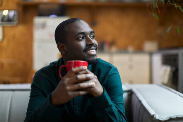 A smiling young man is sitting at home drinking coffee.